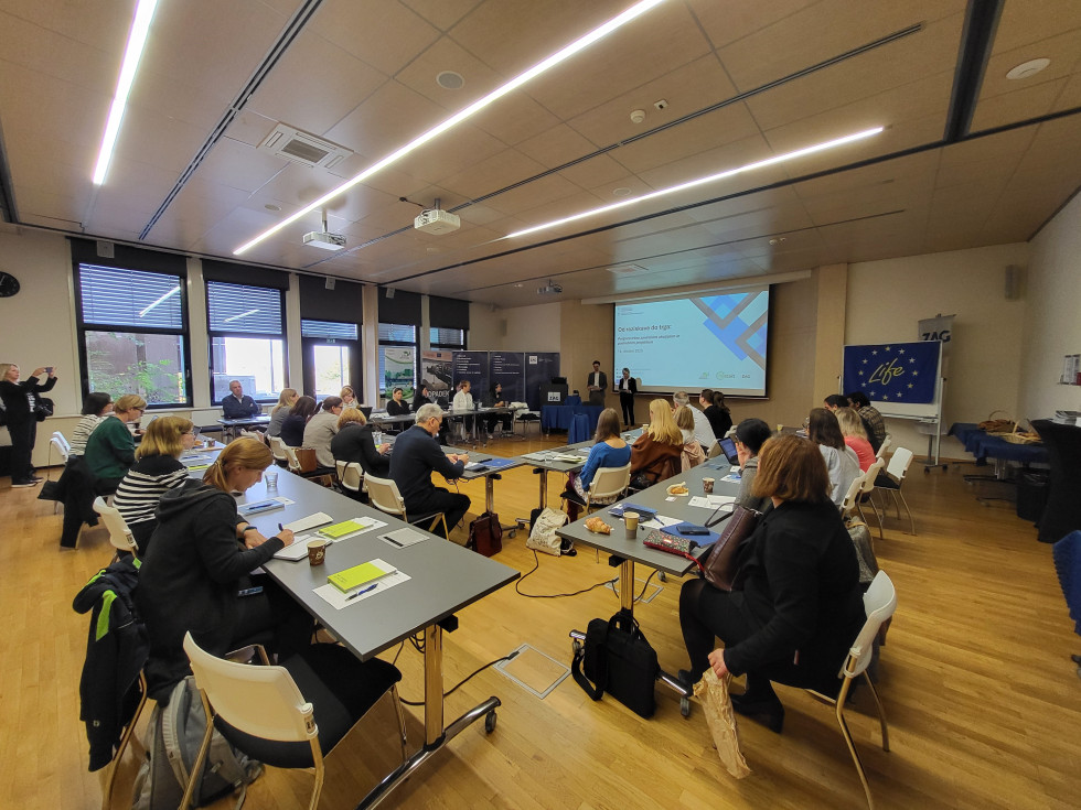 Konferenčna dvorana z mizami in udeleženci, ki sedijo, pred njimi projekcija s predstavitvijo.