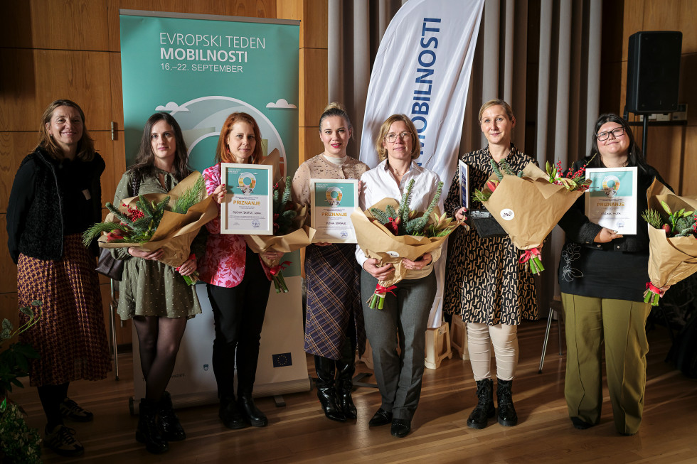 Ljudje v vrsti se fotografirajo, v roki imajo rože in plakete