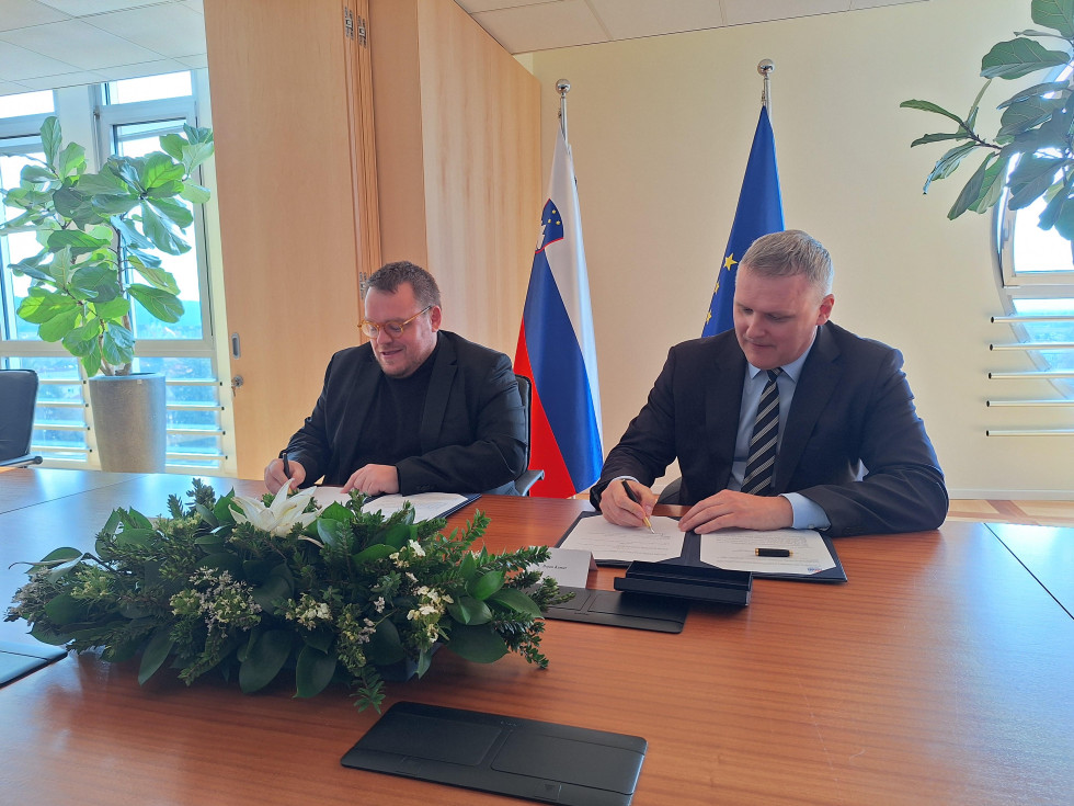 Dva moška za mizo podpisujeta pogodbe, na mizi je zeleno cvetje