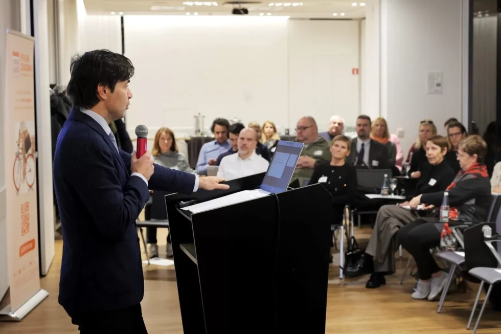moški za govorniškim pultom, pred njim sedi publika