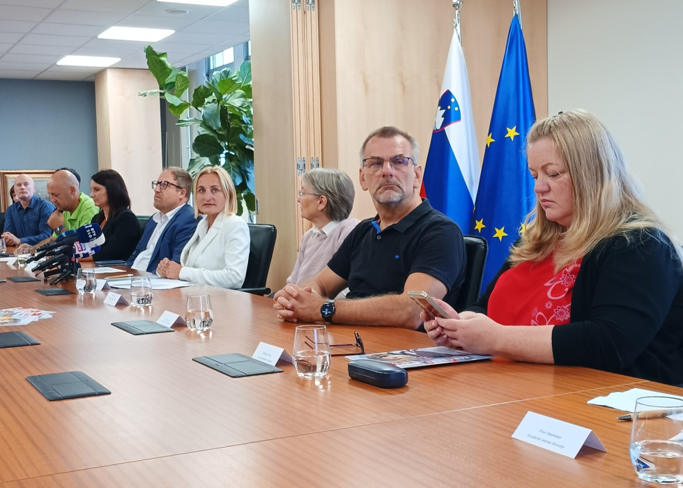 Govorci sedijo za mizo v sejni dvorani, pred njimi mikrofoni slovenskih medijev