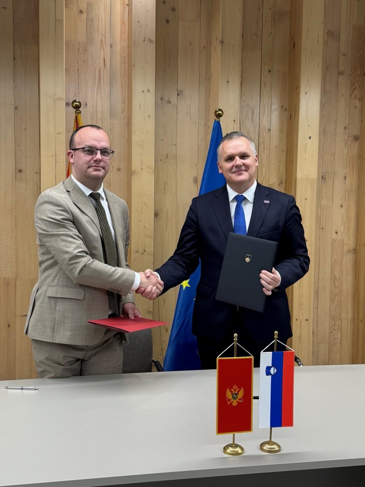 Ministra stojite pred zastavami in se rokujeta. V rokah držita mapi s podpisanima pismoma o nameri.