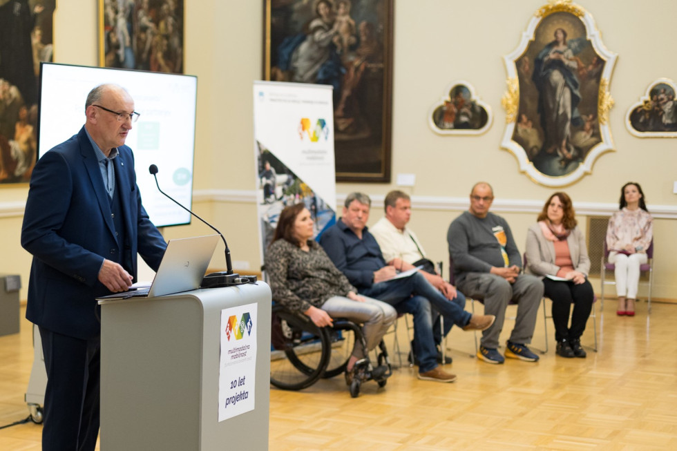 Govorec mag. Roman Rener za govorniškim pultom, v ozadju sedijo predstavniki invalidov in moderatorka