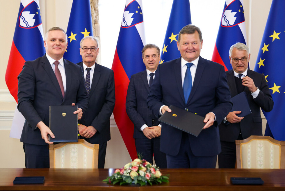 Ministra Kumer in Jevšek stojita in v rokah držita podpisana sporazume. V ozadju stojijo švicarski gospodarski minister in predsednik vlade. za njima zastave Slovenije, in Evropske unije.. 