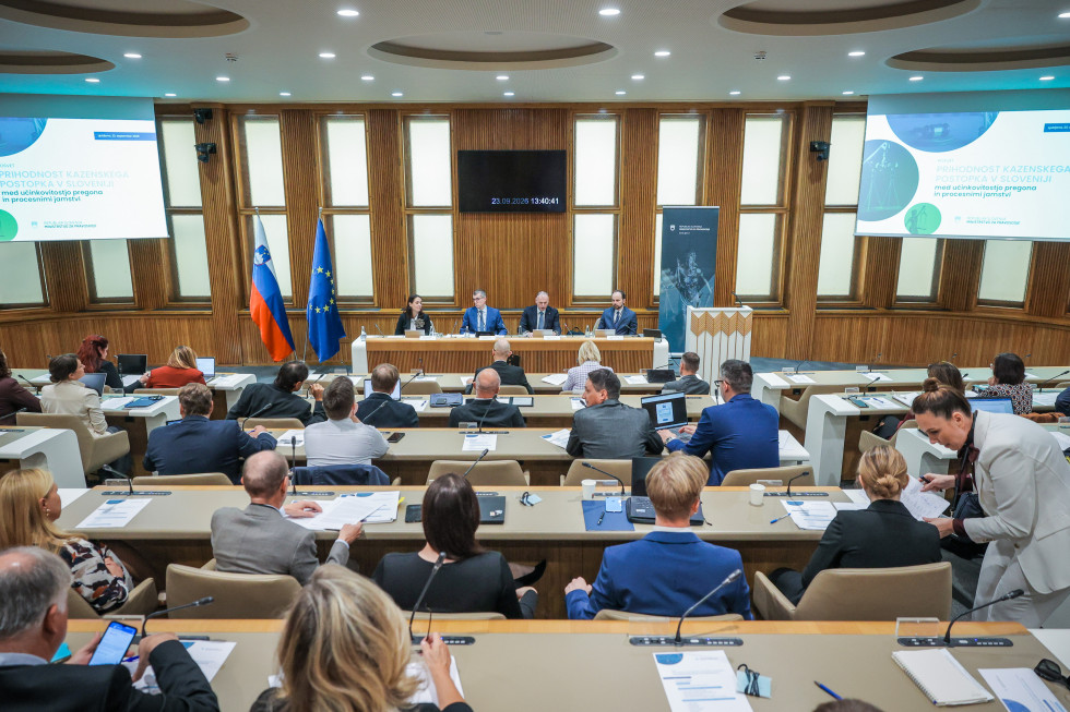 Pogled iz ozadja dvorane Državnega sveta: udeleženci sedijo v klopeh, pred njimi je osrednje omizje s štirimi govorniki, ob strani slovenska in evropska zastava ter zaslona z naslovom posveta.