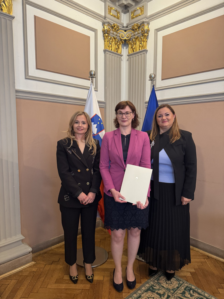 Tri ženske stojijo v okrašeni dvorani s pozlačenimi kapiteli stebrov, v ozadju sta slovenska in evropska zastava. Udeleženka v sredini drži belo mapo z državnim grbom.