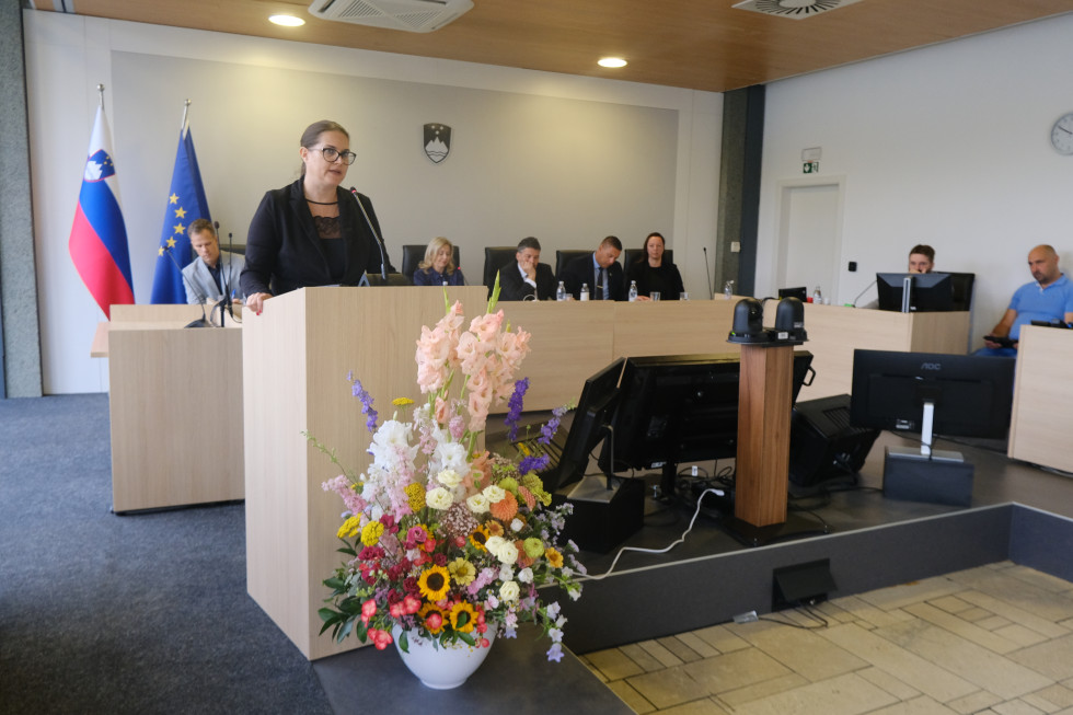 Državna sekretarka mag. Andreja Kokalj stoji za govorniškim pultom in nagovarja udeležence. Pred govorniškim pultom je cvetlični aranžma.