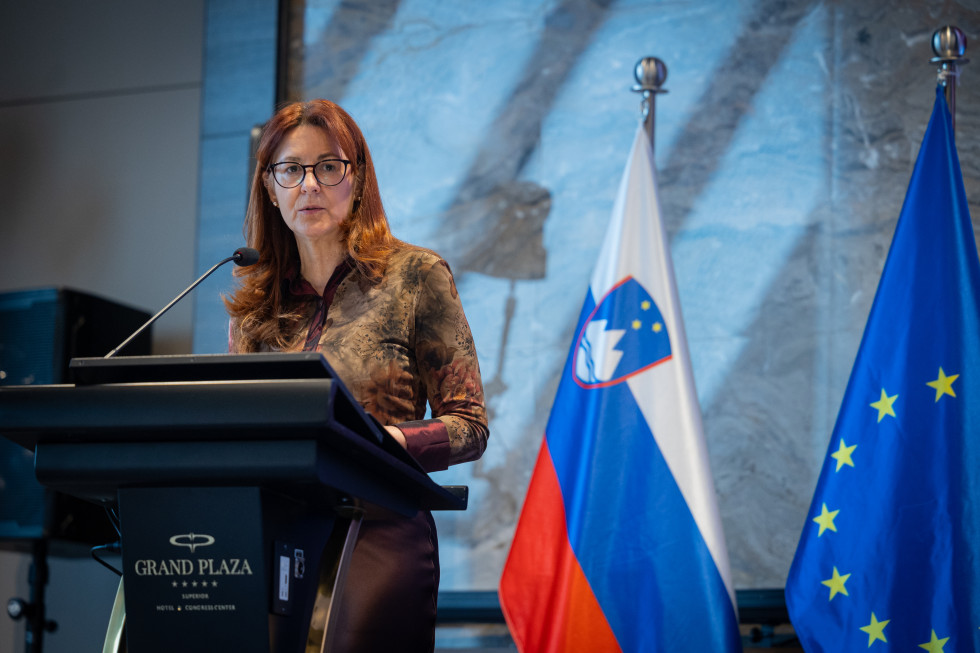 Ministrica Andreja Katič za govornico. V ozadju zastava
