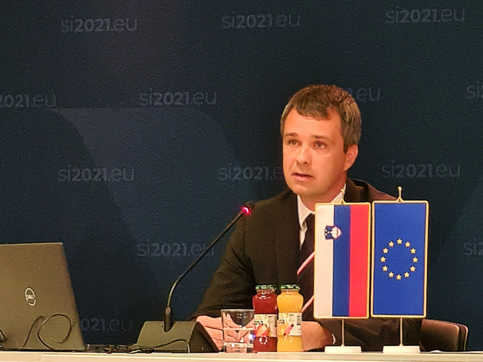 Minister Dikaučič sedi pred panojem predsedovanja in nagovarja člane odbora preko AVK povezave.