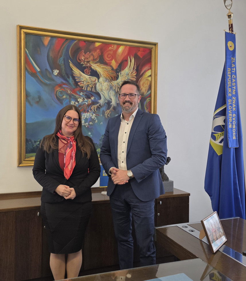 Ministrica za pravosodje in župan Občine Vrhnika stojita v pisarni pred veliko umetniško sliko in zastavo Republike Slovenije ter pozirata za fotografijo.