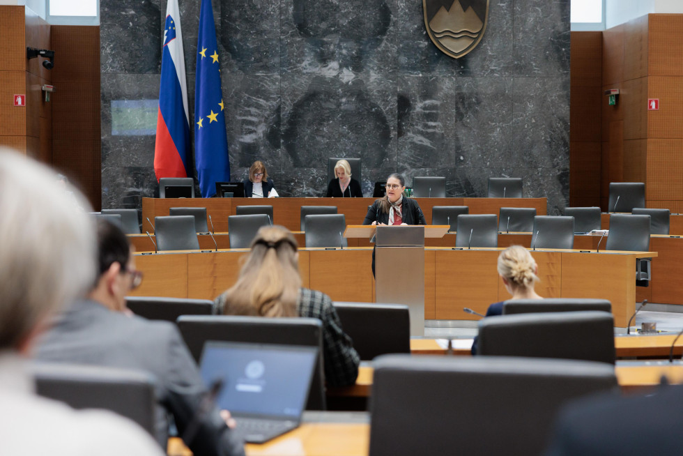 Dvorana v državnem zboru. Ženska stoji za govorniškim pultom in govori. Zadaj slovenska in evropska zastava. Velik grb na steni v ozadju.