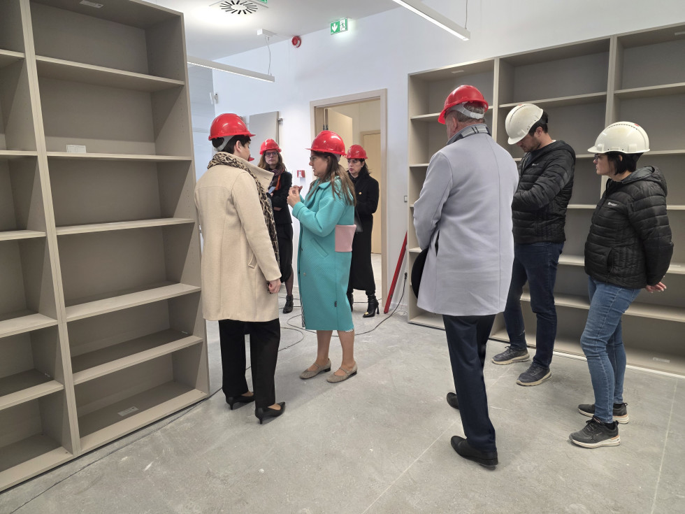 Skupina ljudi z zaščitnimi čeladami stoji v notranjem prostoru in si ogleduje prostor z nameščenimi policami. Ena oseba v sredini razlaga, ostali jo poslušajo. Prostor je svetel, z vgrajenimi omarami in nedokončanimi talnimi površinami.