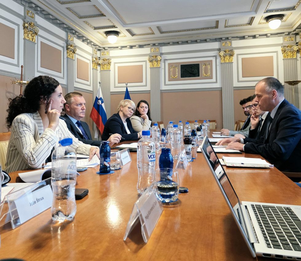 Delegaciji sedita za dolgo sejno mizo na delovnem srečanju ob obisku evropskega komisarja. V ozadju sta slovenska in evropska zastava.