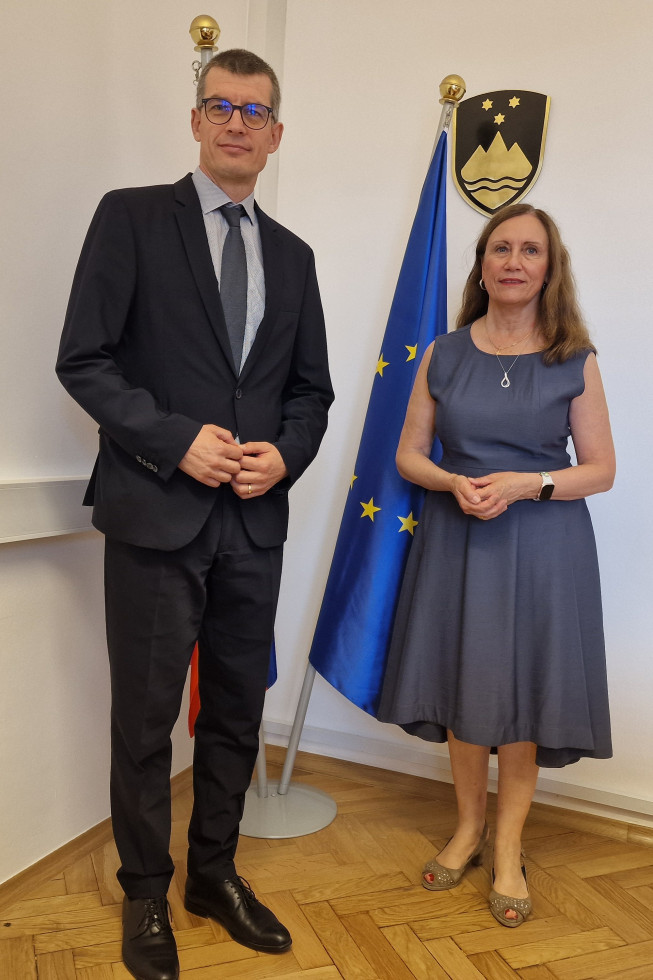 Državni sekretar Gašper Dovžan in generalna sekretarka IHRA Michaela Küchler stojita v prostorih Ministrstva za pravosodje pred slovensko in evropsko zastavo ter državnim grbom.