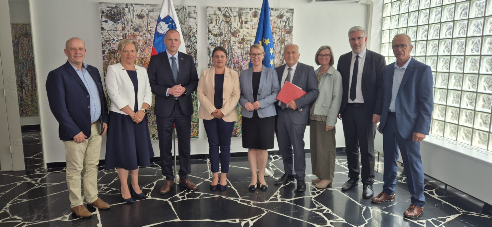 Minister Zupančič, ministrica Lep Šimenko, državna sekretarka, slovenski veleposlanik v Avstriji in predstavniki slovenske narodne skupnosti stojijo na skupinski fotografiji v svetlem prostoru pred slovenskima in evropsko zastavo ter abstraktnimi slikami.