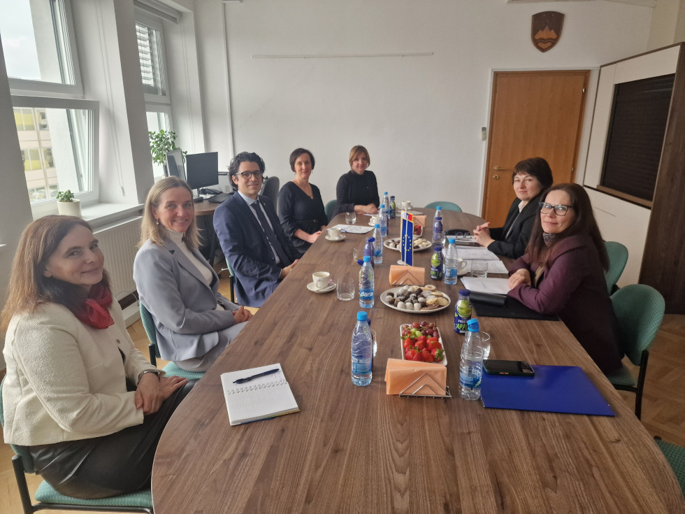 Ženske in en moški sedijo za ovalno leseno mizo. Na mizi papirji, voda in sadje.