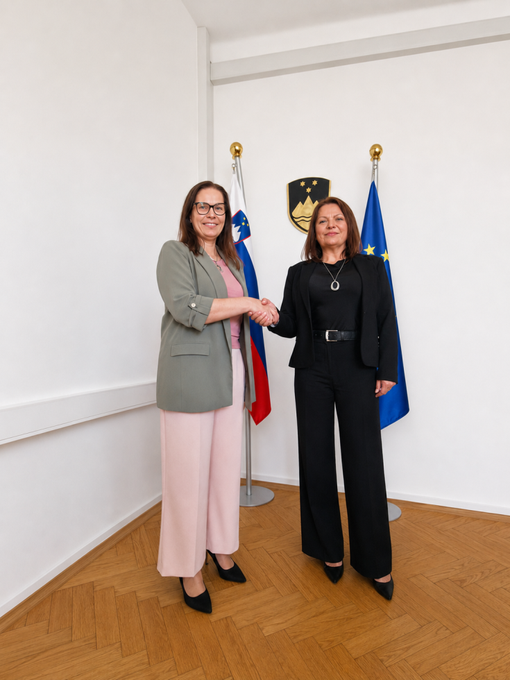 Dve ženski stojita v svetlem protokolarnem prostoru in si rokujeta pred slovensko ter evropsko zastavo in državnim grbom. Ena je oblečena v svetel suknjič in rožnate hlače, druga v črn poslovni kostim. Fotografija deluje uradno in slovesno.