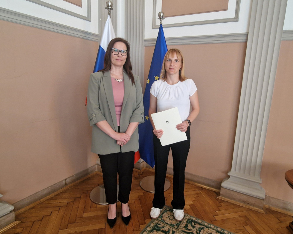 Državna sekretarka dr. Maja Čarni Pretnar in sodna izvedenka Katarina Walland po zaprisegi na Ministrstvu za pravosodje, pred slovensko in evropsko zastavo.