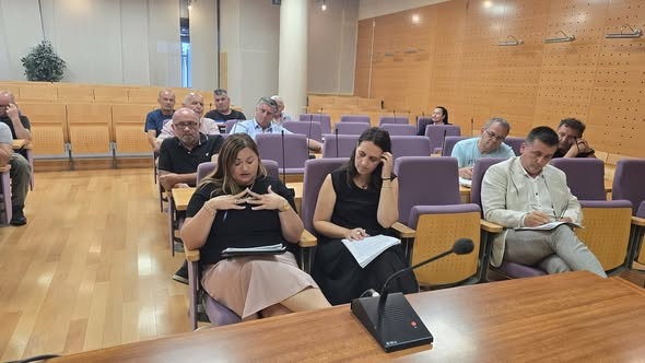 Udeleženci posveta sedijo v sejni dvorani; v ospredju tri osebe spremljajo gradivo in si zapisujejo, pred njimi je konferenčni mikrofon.