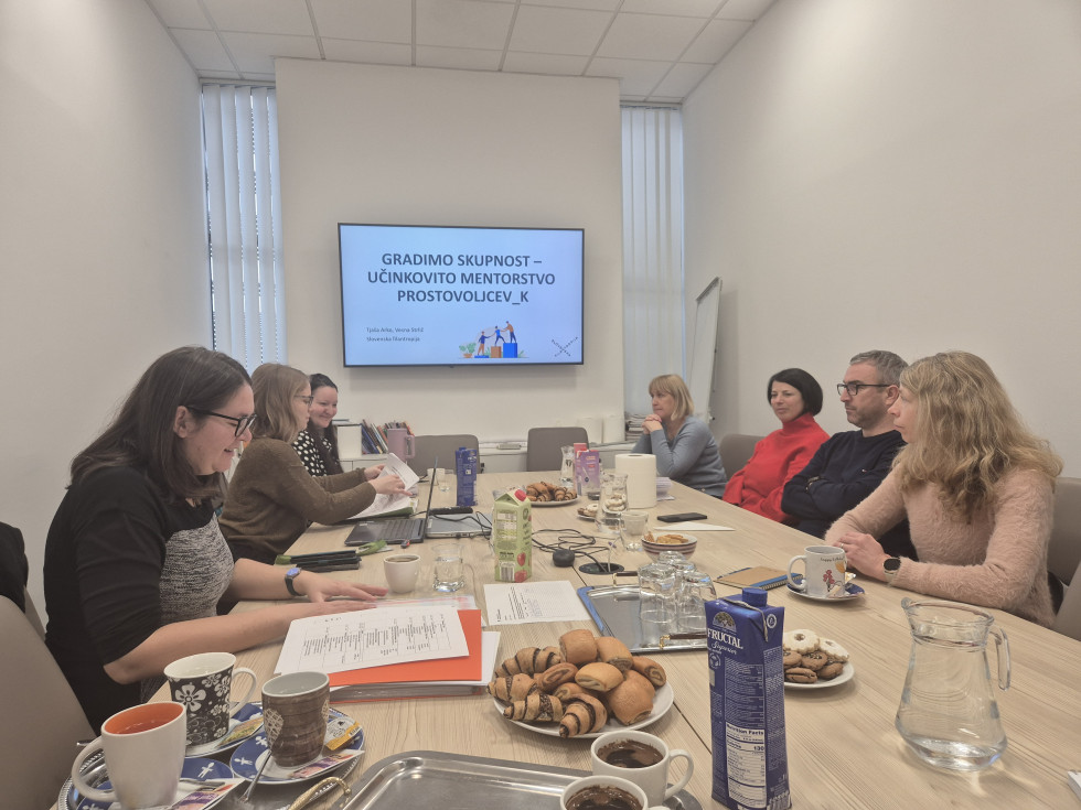 Koordinatorji sedijo za mizo, v ozadju veliki ekran