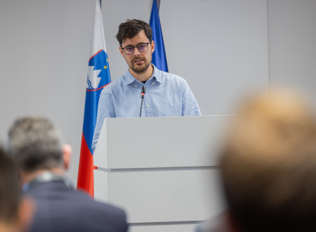 Državni sekretar pred govorniškim pultom, v ozadju zastave