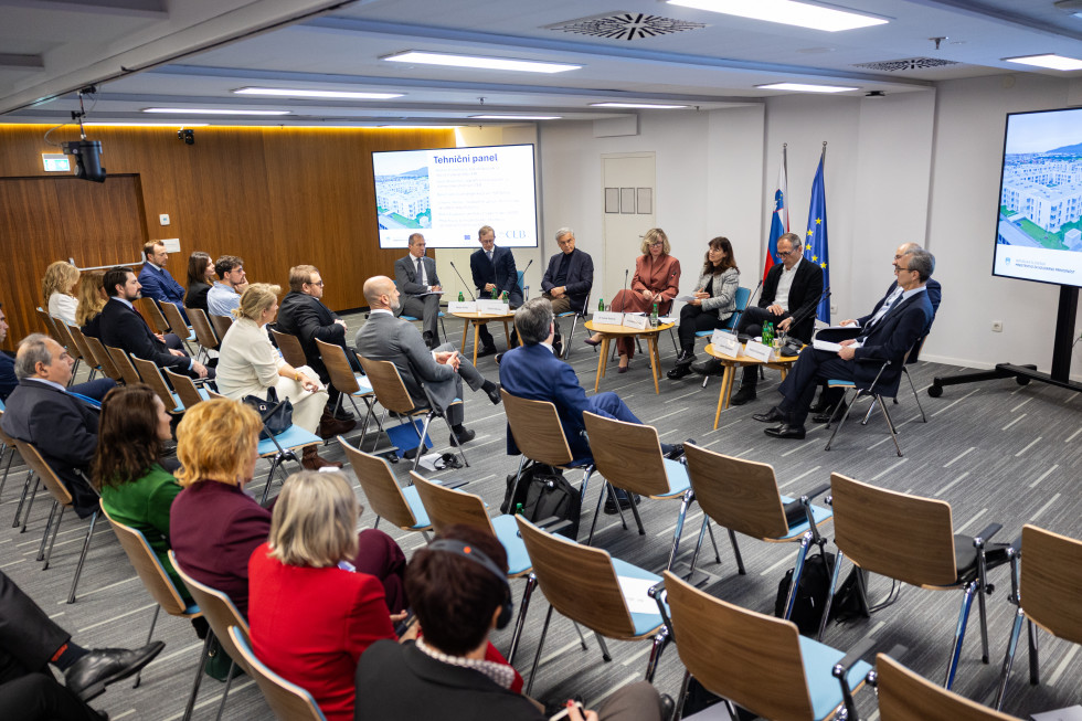 Gostje panela v ozadju, spredaj prazni stoli in udeleženci konference