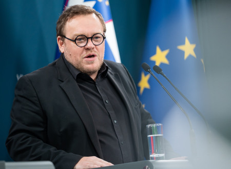 Minister stoji za govorniškim pultom, zadaj zastave