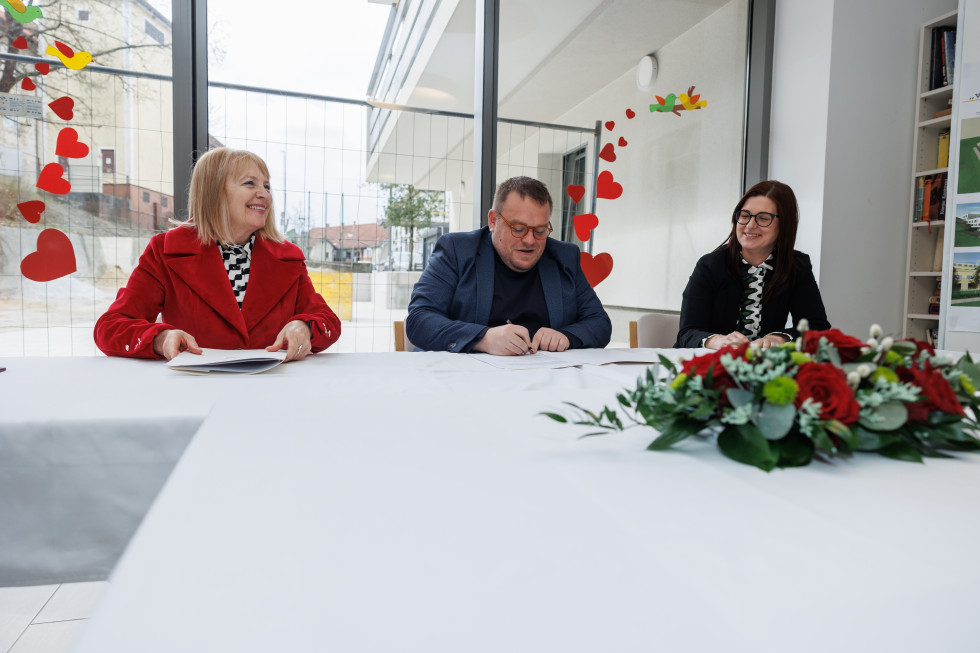 Podpis pogodbe. Minister podpisuje pogodbo, zraven njega sedita dve ženski. Na mizi lep šopek.