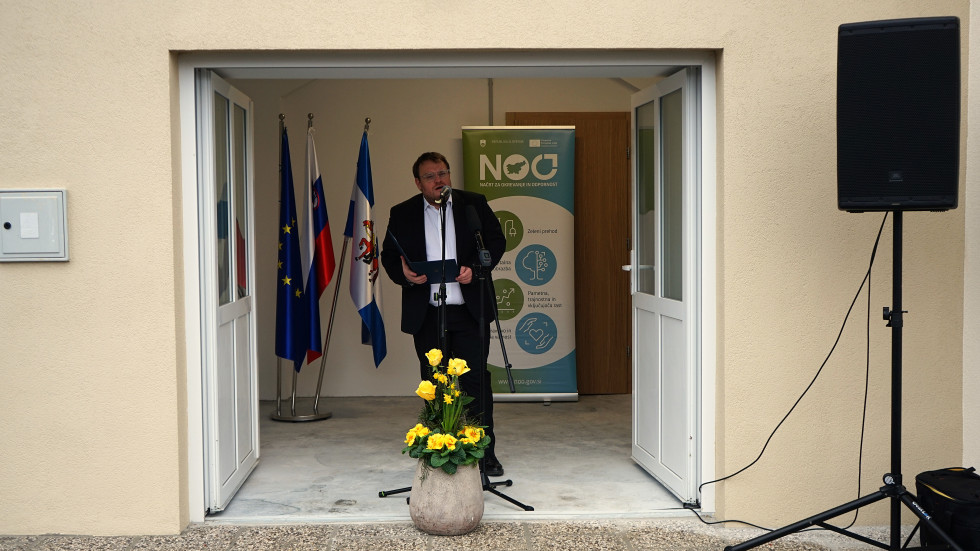 Minister stoji pred mikrofonom, v ozadju zastave in pano Načrta za okrevanje in odpornost