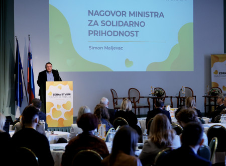 Minister ima nagovor. Stoji za govorniškim pultom. Za njim na velikem platnu prezentacija. Občinstvo sedi in spremlja nastop.