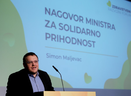 Minister ima nagovor. Stoji za govorniškim pultom. Zadaj na velikem platnu je prezentacija.