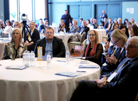 Minister sedi za mizo z ministrico za zdravje Valentino Prevolnik Rupnik. Za okroglimi mizami sedijo udeleženci in spremljajo nastop.