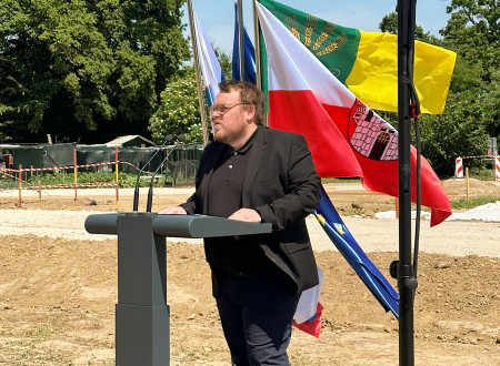 Minister stoji za govorniškim pultom, za njim zastave, zadaj gradbišče