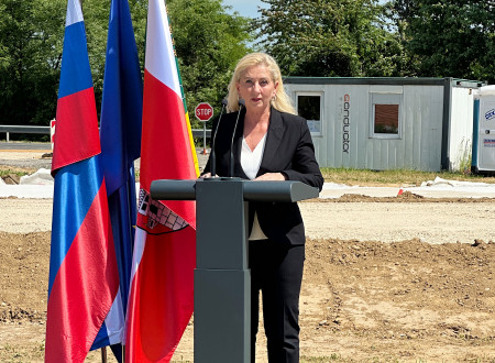 Direktorica stoji za govorniškim pultom, levo od nje zastave, zadaj gradbišče