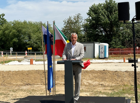 Župan stoji za govorniškim pultom, levo od njega zastave, zadaj gradbišče