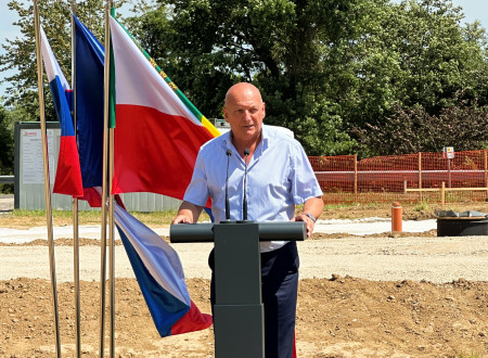 Župan stoji za govorniškim pultom, levo od njega zastave, zadaj gradbišče