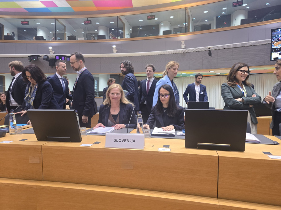 Državna sekretarka sedi v dvorani. Pred njo tablica z napisom: Slovenija. 