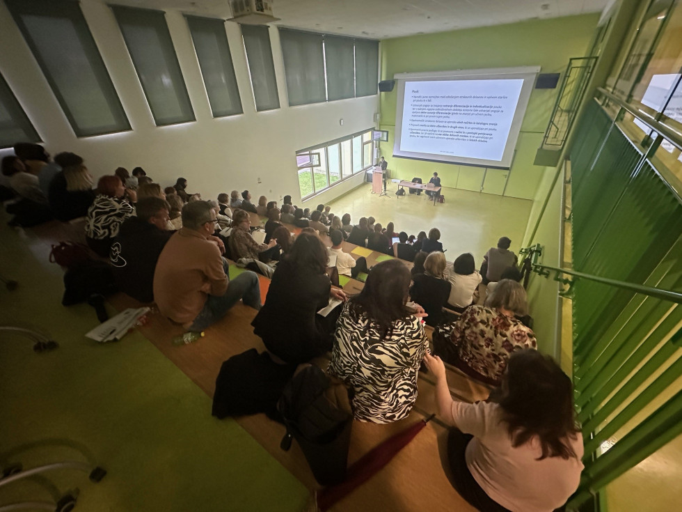 Slika velike predavalnice v osnovni šoli, gledano iz zadnje vrste udeležencev. Vodja izza pulta nagovarja udeležence, za njim je velika projekcija. 