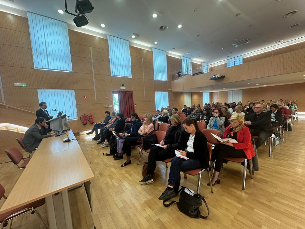 Slika prikazuje udeležence posveta, ki sedijo v dvorani in poslušajo vodjo delovne skupine, ki predstavlja nacionalni program stoje izza pulta.