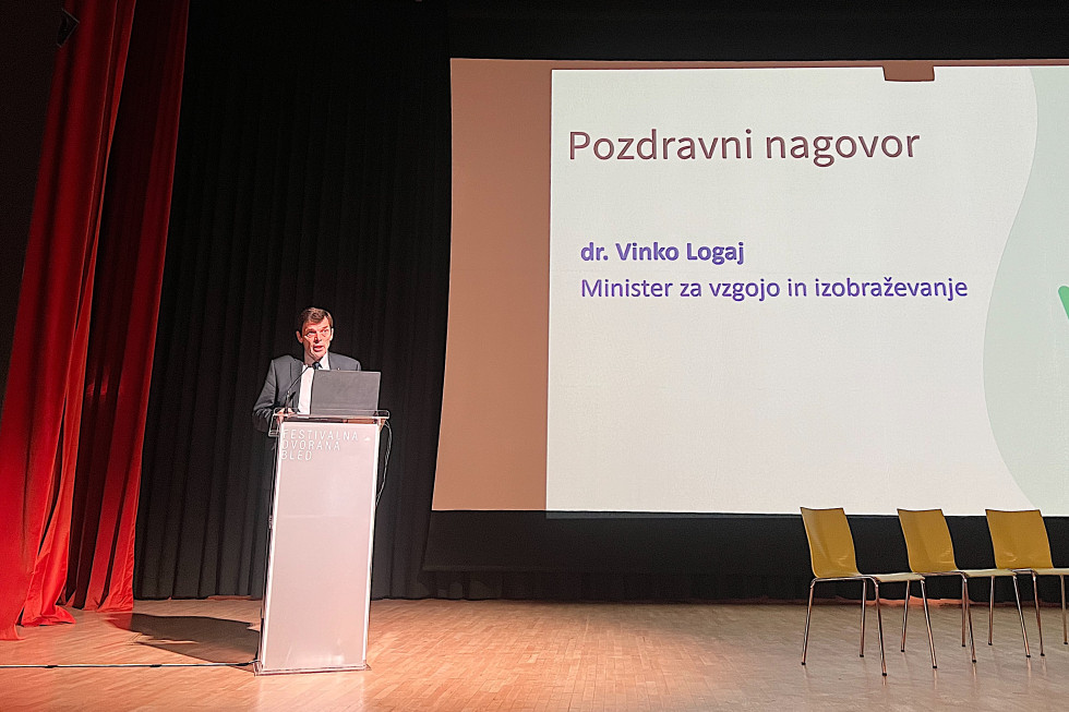Minister stoji za govorniškim pultom na odru dvorane in nagovarja udeležence konference, ob njem je velik zaslon z napisom: Pozdravni nagovor dr. Vinko Logaj minister za vzgojo in izobraževanje.