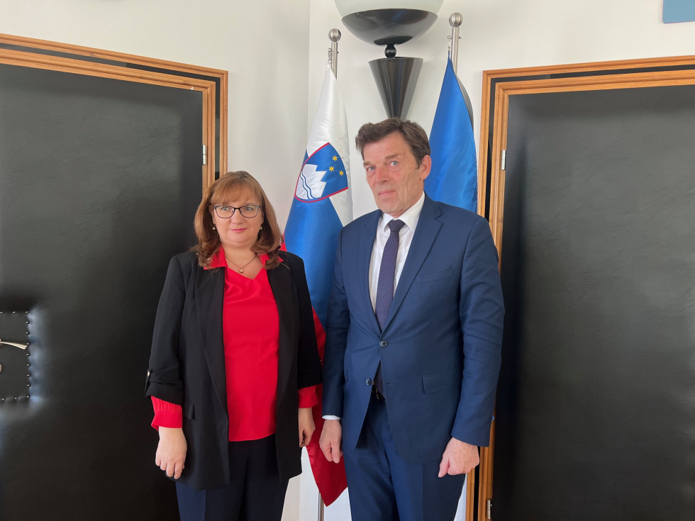 varuhinja in minister v pisarni pred zastavami