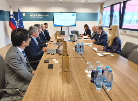 Delegaciji na uradnem sestanku za mizo, na zaslonu predstavitev o raziskovalnih dejavnostih KEK.