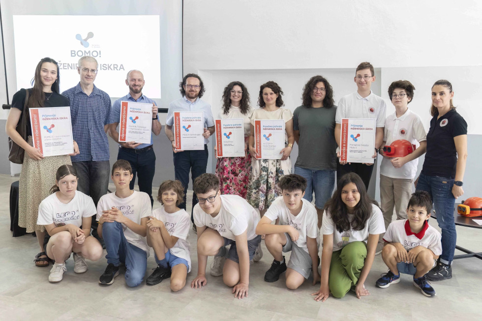 Skupinska fotografija nagrajencev in prejemnikov priznanj, ki stojijo in klečijo pred projekcijskim platnom; več oseb drži uokvirjena priznanja.