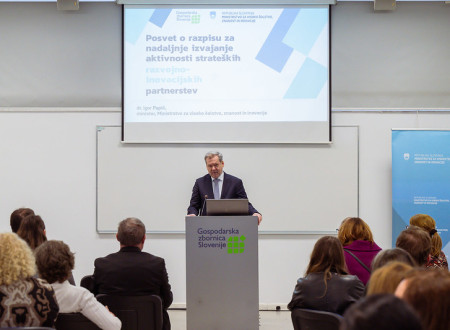 Moški stoji za govornico in govori. Zadaj je predstavitev in napis: Posvet o razpisu za nadaljnje izvajanje strateških razvojnih partnerjev.