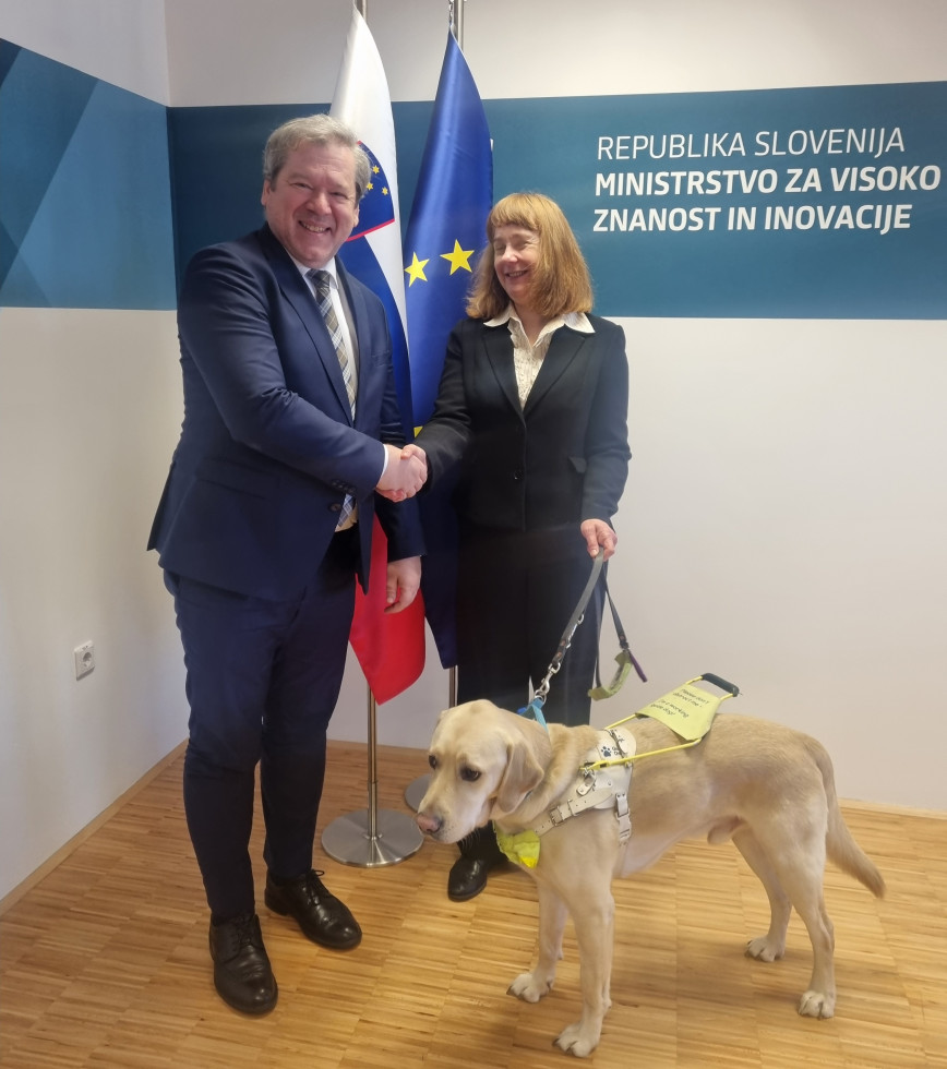 Minister Papič med rokovanje z veleposlanico Victorio Harrison. V ospredju pes vodnik, v ozadju slovenska in evropska zastava.