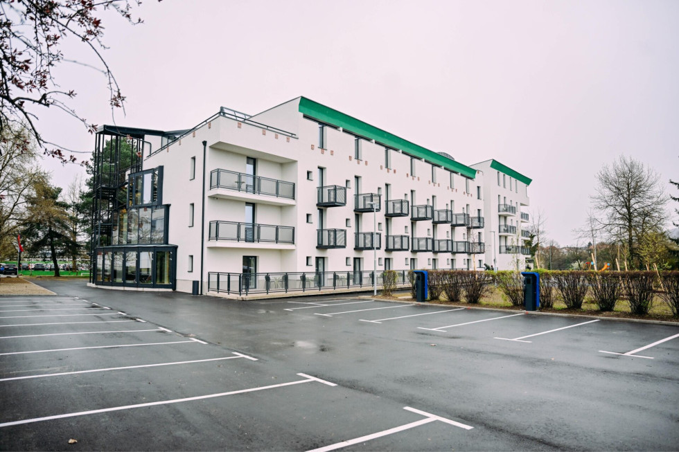 Izgled prenovljenega doma. Sodobna večetažna stavba z belimi fasadami, balkoni in steklenim prizidkom, obdana z asfaltiranim parkiriščem in drevjem v ozadju.