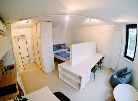 Svetel enosobni prostor z majhno kuhinjo, jedilnim pultom, delovno mizo in posteljo, opremljen v svetlih barvah.