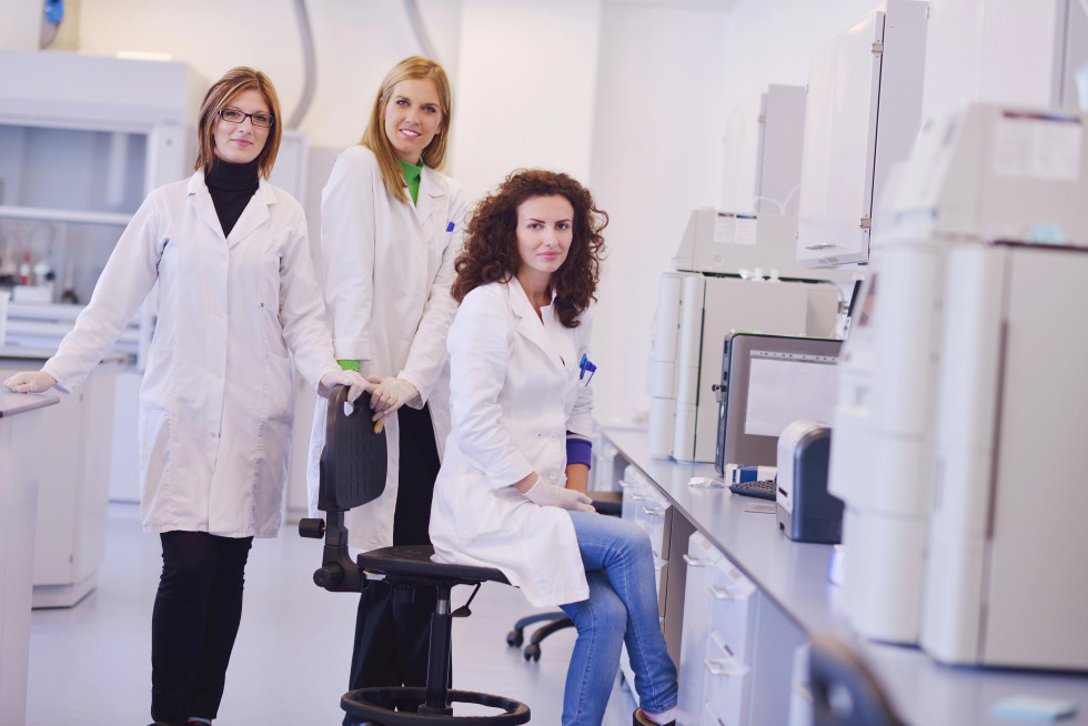 Znanstvenice v belem laboratorijskem plašču v laboratoriju. V ozadju so laboratorijske naprave in računalniki.