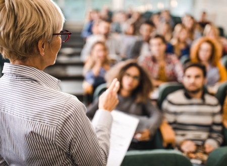 Profesorica med predavanjem, v ozadju študentke in študenti v predavalnici.