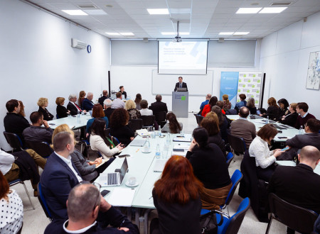 Moški stoji za govornico in govori polni sobi. Ljudje sedijo za mizami in opazujejo govorca.